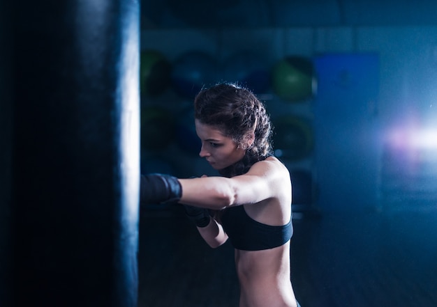 重いサンドバッグでトレーニング中のボクシンググローブを身に着けている若い戦闘機ボクサーフィットの女の子