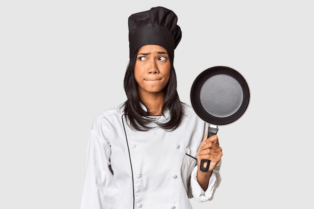 Photo young filipina chef holding pan by handle confused feels doubtful and unsure