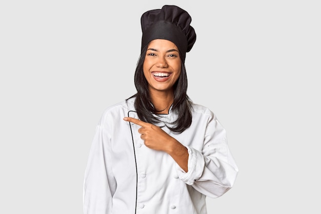 Photo young filipina chef with cooking hat in studio smiling and pointing aside showing something