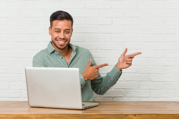 Il giovane uomo filippino seduto a lavorare con il suo laptop eccitato indicando con gli indici di distanza