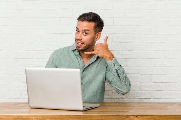 Young filipino man sitting working with his laptop showing a mobile phone call gesture with fingers