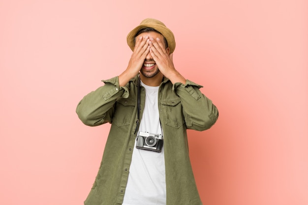 Young filipino traveler man covers eyes with hands, smiles broadly waiting for a surprise.