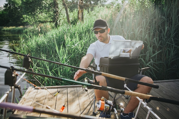 Giovane pescatore che pesca sul lago o sul fiume. ragazzo adulto seduto sulla sedia pieghevole e allungando la mano a una delle aste. prepararsi per la pesca. regolazione dell'attrezzatura per la caccia in acqua.