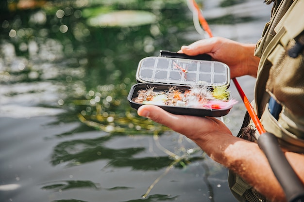 湖や川で釣りをする若い漁師。開いた箱で魚を捕まえるためにいくつかのプラスチック製の人工ルアーを持っている男の写真をクローズアップ。水中で一人で立ち、釣り竿を持ってください。