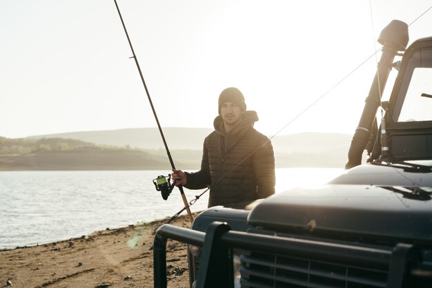 Young fisherman standing near his car and holding fishing equipment