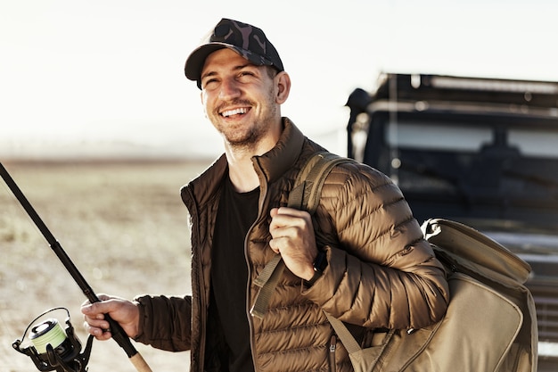 Young fisherman standing near his car and holding fishing equipment