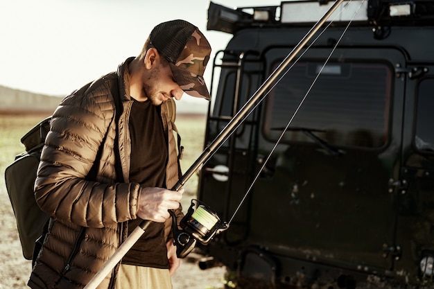 Young fisherman standing near his car and holding fishing equipment