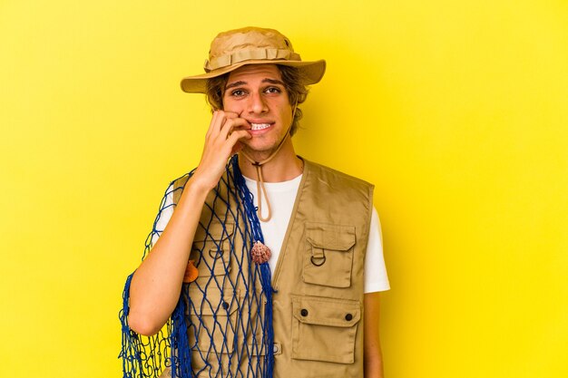 Young fisherman with makeup holding a net isolated on yellow background  biting fingernails, nervous and very anxious.