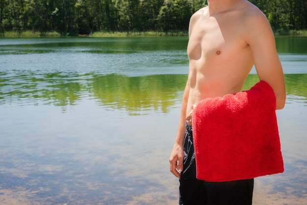 Giovane e in forma, uomo atletico in piedi sulla riva del lago senza camicia mentre tiene un asciugamano rosso. nuoto, attività estive nella natura