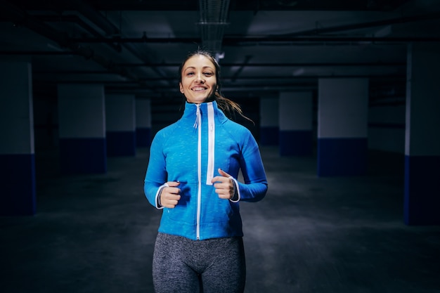 Young fit caucasian sportswoman in tracksuit running fast in garage.