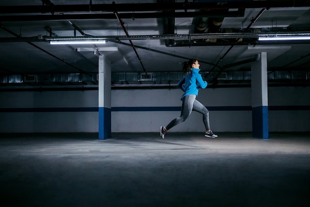Young fit caucasian sportswoman in tracksuit running fast in garage.