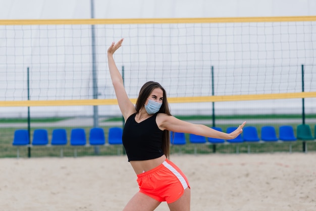 Молодая женщина в спортивной одежде и защитной маске от коронавируса на красной дорожке и волейбольной площадке