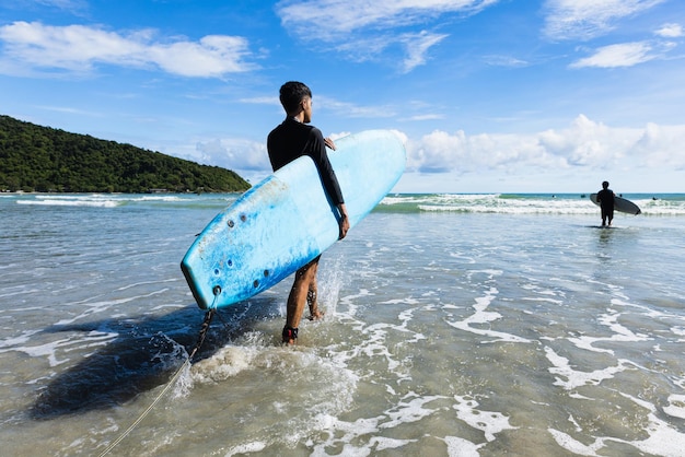 Foto ragazzo adolescente in forma e corpo sodo che cammina in mare tenendo la tavola morbida pronta per gli sport estremi che praticano il surf sulle vacanze al mare nei cieli azzurri della giornata limpida