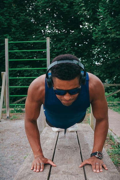 Young fit man with sunglasses and a blue shirt doing push-ups in a park while listening to music