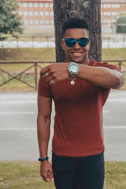 Young fit man with sunglasses and a cool hairstyle showing his watch near the tree