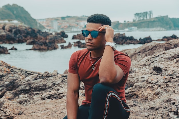 Young fit man with sunglasses sitting on the rock near the calm sea during daytime