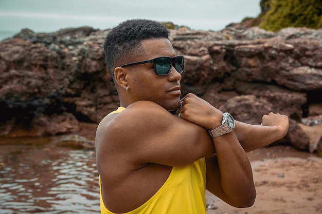 Young fit man with sunglasses stretching his arms with the calm ocean in the background