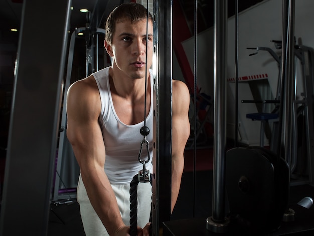 Молодой спортсмен в белой футболке делает упражнения на трицепс на тренажере для кросса в тренажерном зале