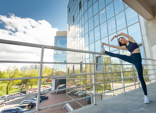 Young fit woman stretches her leg from the railing on the balcony of urban outhouse against the backdrop of building Healthy lifestyle