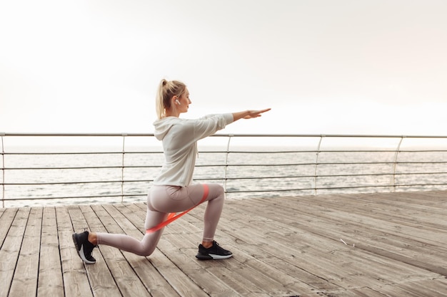 Young fit woman trains legs with fitness rubber bands on the beach. Healthy lifestyle