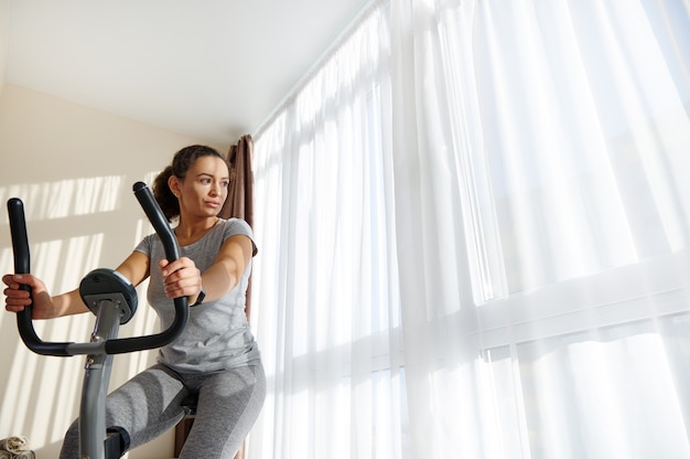 Young fit woman using stationary bike for cardio workout at home