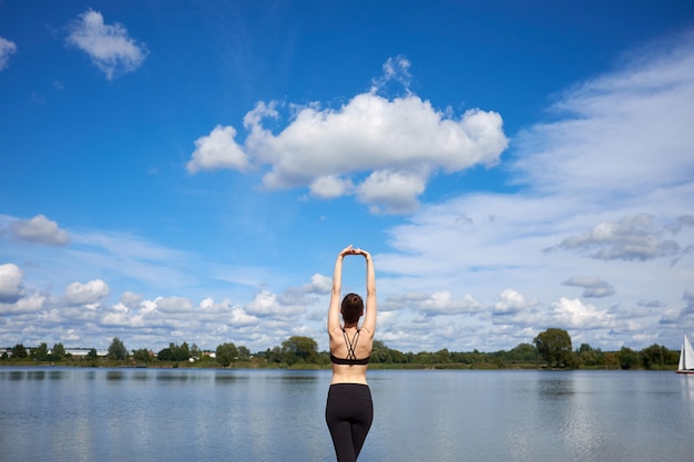 Young fit woman wearing black stylish sportswear stretching near lake outdoor.