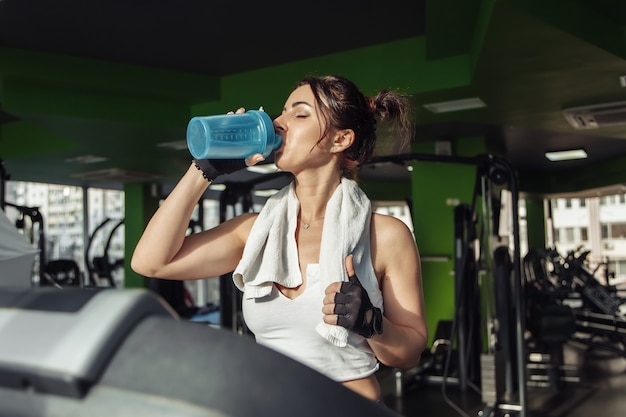 肩にタオルを持った若いフィットの女性は、トレッドミルで水を飲みます。減量の概念、有酸素運動