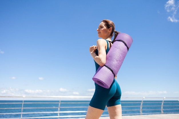 Young fit woman with yoga mat stands on the embankment terrace on a bright sunny day