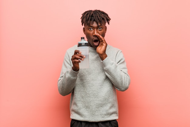 Young fitness black man holding a water bottle shouting excited to front.