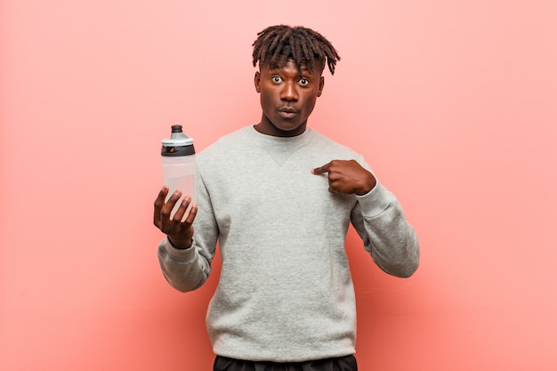 Young fitness black man holding a water bottle surprised pointing at himself, smiling broadly.