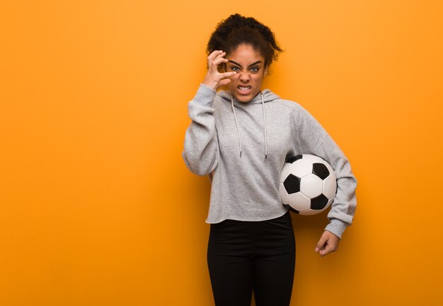 Young fitness black woman angry and upset. Holding a soccer ball.