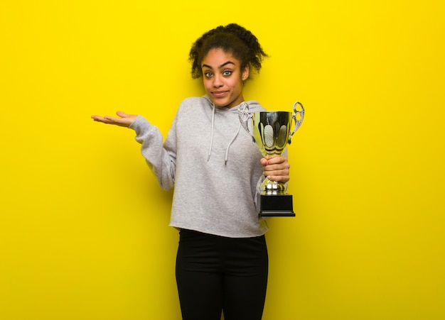 Photo young fitness black woman doubting and shrugging shoulders. holding a trophy.