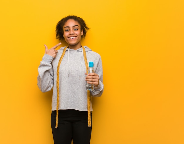 Young fitness black woman smiles, pointing mouth.Holding a water bottle.