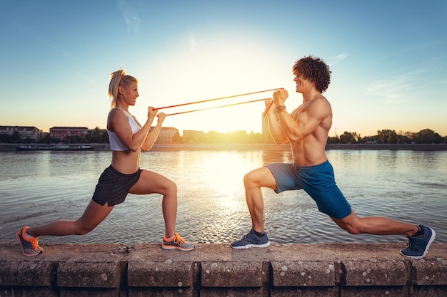 Young fitness couple doing workout with rubber band by the river in a sunset.