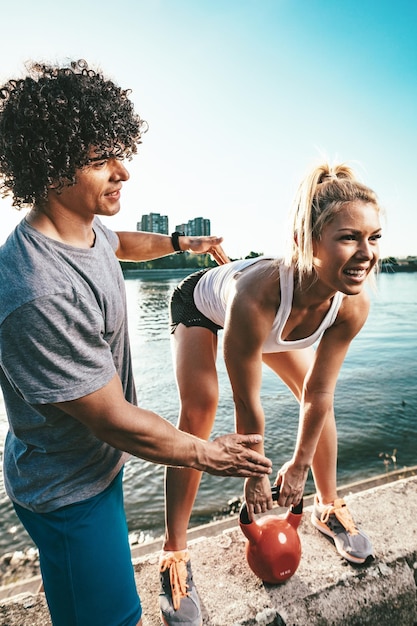 Young fitness couple is doing workout with kettlebell on the wall  by the river in a sunset. The woman is crouching and holding kettlebell, and the man supports her.