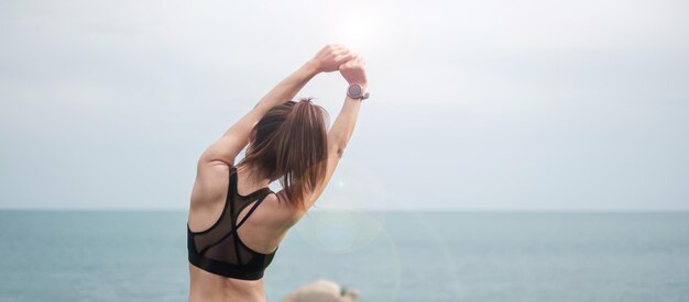 Young fitness female in sportswear stretching body against ocean view, healthy woman exercise in morning. Workout, wellness and work life balance concepts