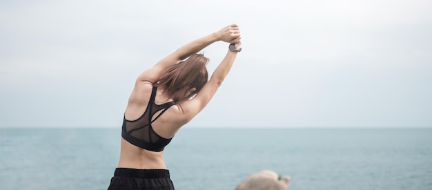 Young fitness female in sportswear stretching body against ocean view, healthy woman exercise in morning. Workout, wellness and work life balance concepts