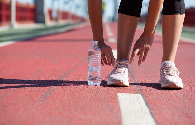 Giovane ragazza di forma fisica che lega i lacci delle scarpe sulla pista da corsa in una giornata di sole. spazio per il testo