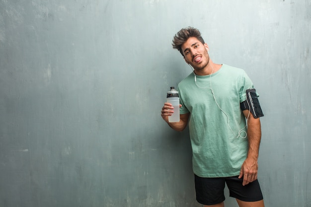 Young fitness man against a grunge wall expression of confidence and emotion, fun and friendly, showing tongue as a sign of play or fun. Wearing an armband with phone.