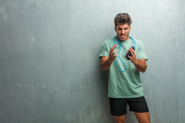 Young fitness man against a grunge wall very angry and upset, very tense, screaming furious, negative and crazy. Holding a jump rope.