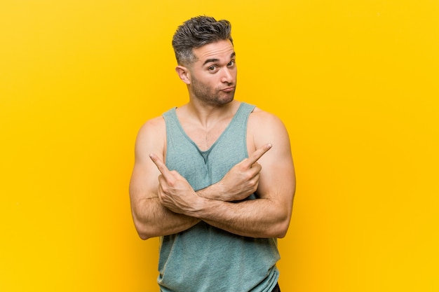 Young fitness man against a yellow wall points sideways, is trying to choose between two options.