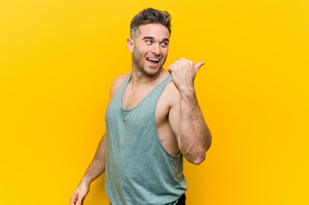 Young fitness man against a yellow wall points with thumb finger away