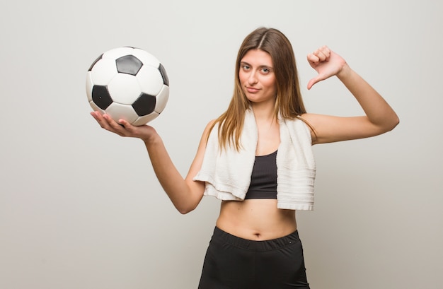 Young fitness russian woman pointing fingers, example to follow. Holding a soccer ball.