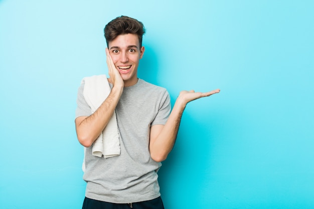 Young fitness teenager man holds copy space on a palm, keep hand over cheek. Amazed and delighted.