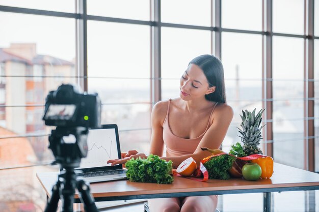 Young fitness vlogger doing video indoors by sitting near table with healthy food.