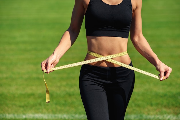 Young fitness woman in black sportswear with perfect waist with a measuring tape in hands