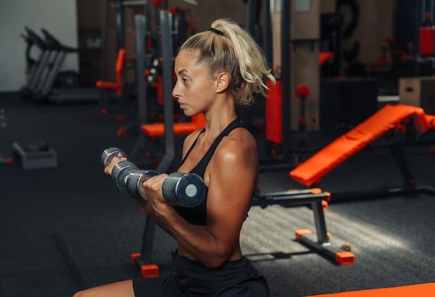 Young fitness woman doing dumbbell lifts for biceps while sitting on a bench in the gym. Training concept with free weights.