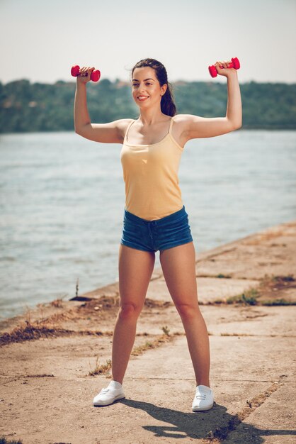 川のそばでダンベルで運動をしている若いフィットネス女性。