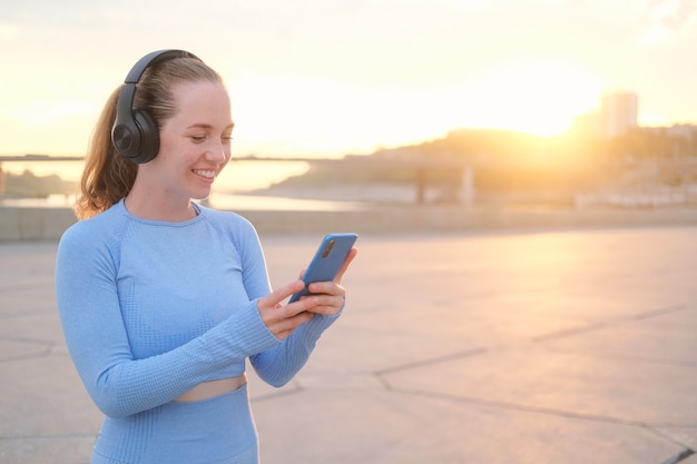 若いフィットネス女性が街を走り終える。携帯電話を持ちながら、ヘッドホンで音楽をお楽しみください。日没。健康的な生活様式。自由。屋外トレーニング。高品質の写真