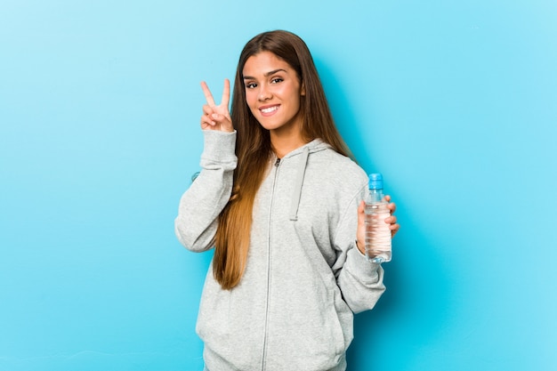 Молодая женщина фитнеса держа бутылку с водой показывая знак победы и широко усмехаясь.